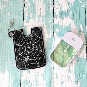May include: A black glittery fabric holder featuring a white stitched spiderweb and spider design with a silver metal keychain clip, paired with a white rectangular hand sanitizer spray bottle containing green liquid labeled Touchland Power Mist 1 fl oz.