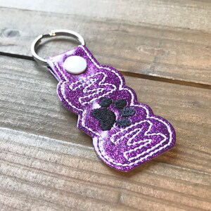 May include: Purple glitter keychain with a white stitched border and the word "Mom" with paw prints in the center.