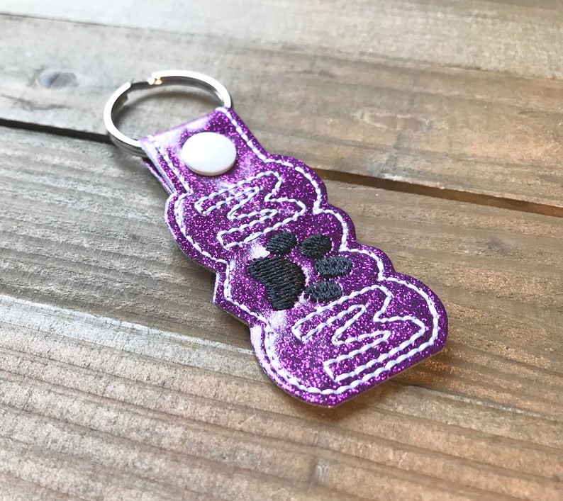 May include: Purple glitter keychain with a white stitched border and the word "Mom" with paw prints in the center.