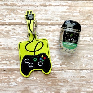 May include: A green and black video game controller keychain with a black cord and plug. The keychain is next to a small bottle of hand sanitizer with the text "Stress Relief Eucalyptus Spearmint with Essential Oils PocketBac 1 fl oz/29 ml" on the label.