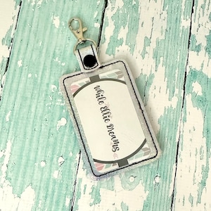 May include: A white glitter key chain with a black strap and a silver clip. The key chain has a rectangular tag with a white background and black text that reads "while ellie dreams".
