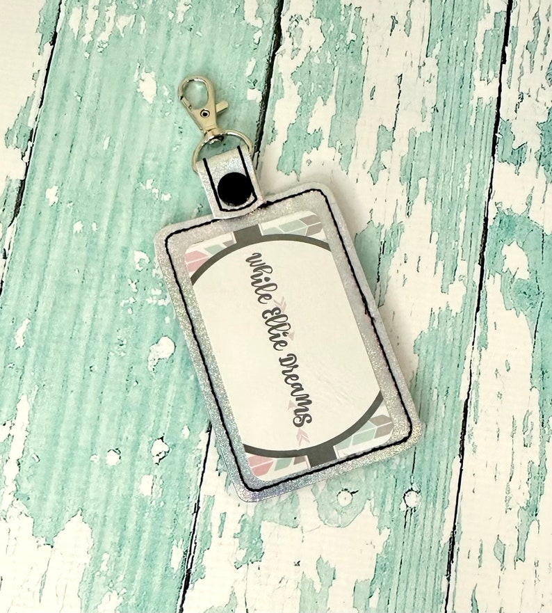 May include: A white glitter key chain with a black strap and a silver clip. The key chain has a rectangular tag with a white background and black text that reads "while ellie dreams".