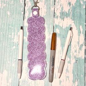 May include: A purple glitter keychain with a scalloped edge, a silver clasp, and two white pens with gray grips, and a gold Sharpie marker. The items are arranged on a distressed, light blue wooden surface.
