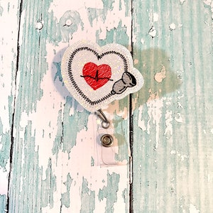 May include: White heart-shaped badge holder with a red heart and EKG line, a stethoscope, and a clear retractable badge reel.