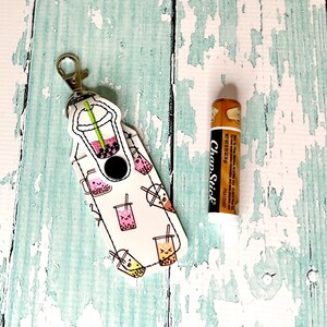 May include: A white fabric keychain with a bubble tea design and a black snap closure. The keychain has a silver metal ring and a silver metal clip. A brown lip balm tube with the text "ChapStick" is also shown.