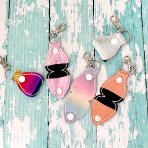 May include: Five keychains with different designs. Each keychain has a metal ring and a fabric pouch with a snap closure. The pouches are in various colours and patterns, including rainbow, pink, and peach.