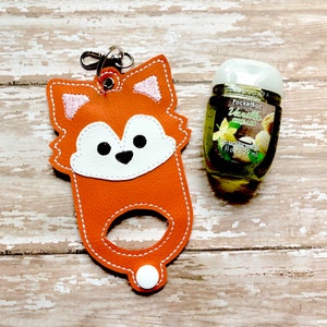 May include: Orange fox-shaped leather hand sanitizer holder with a white face and a white button. The holder has a silver keychain attached. A clear plastic bottle of PocketBac Vanilla Cream hand sanitizer is next to the holder.