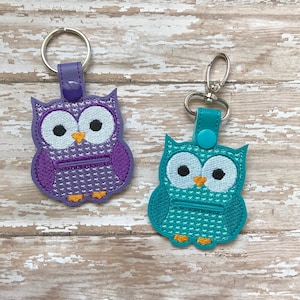 May include: Two owl-shaped keychains with zippered pockets. The owl on the left is purple with white eyes and a yellow beak. The owl on the right is teal with white eyes and a yellow beak.