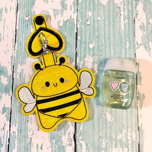 Peut inclure: Un porte-gel hydroalcoolique en forme d'abeille jaune et noir avec un porte-clés en forme de cœur. Le porte-gel est en tissu et a une finition pailletée. Une petite bouteille de gel hydroalcoolique est représentée à côté du porte-gel.