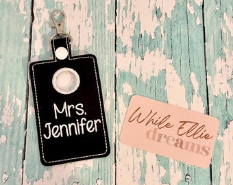 May include: A black rectangular keychain holder with a silver metal lobster clasp and a white snap button. The holder features a circular cutout and white embroidered text that reads Mrs. Jennifer. Beside it is a light pink card with the text While Ellie dreams.