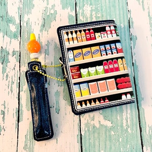 May include: A blue denim pencil case with a yellow and orange plastic pencil topper. The pencil case has a zipper closure and a small loop for attaching a keychain. The pencil case is in front of a colorful fabric wallet with a design of a grocery store shelf filled with various food items.
