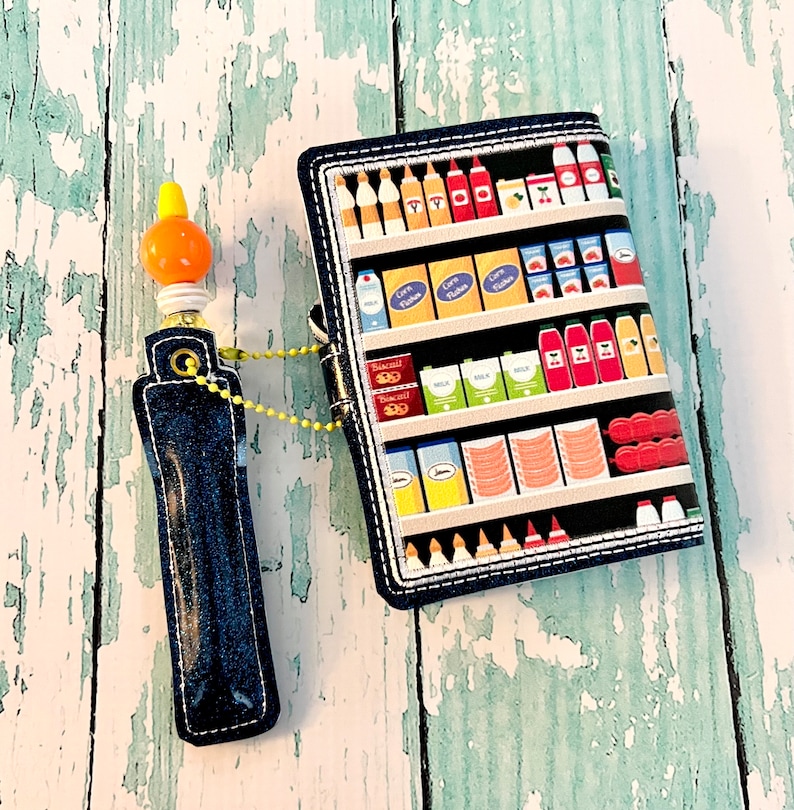 May include: A blue denim pencil case with a yellow and orange plastic pencil topper. The pencil case has a zipper closure and a small loop for attaching a keychain. The pencil case is in front of a colorful fabric wallet with a design of a grocery store shelf filled with various food items.