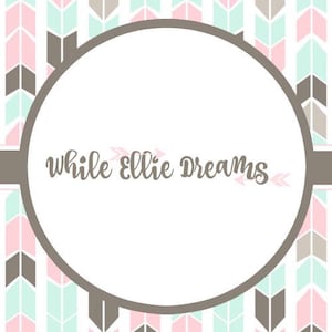May include: A white circle with the text "while ellie dreams" in brown cursive lettering. The circle is surrounded by a pattern of pink and green arrows on a white background.