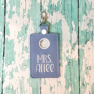 May include: A light blue key chain with a white button and a clear plastic window. The keychain has the text "MRS. Allee" embroidered in white thread.