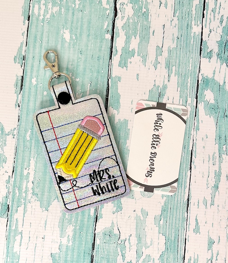 May include: A silver glitter keychain with a yellow pencil and the text "Mrs. White" on a white background. The keychain has a silver metal clasp.
