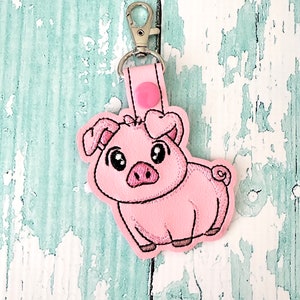 Pode incluir: Chaveiro de porco rosa com um coração na cabeça. O porco é bordado em um tecido rosa com costuras pretas. O chaveiro tem um fecho de metal prateado.