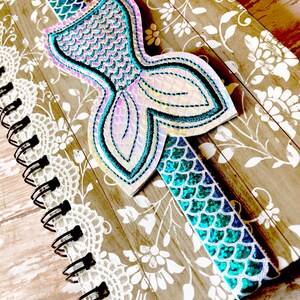 Puede incluir: Un cuaderno con un marcap&aacute;ginas en forma de cola de sirena. El marcap&aacute;ginas est&aacute; hecho de tela iridiscente con un esquema de color azul y verde. El cuaderno tiene un dise&ntilde;o floral marr&oacute;n y blanco.