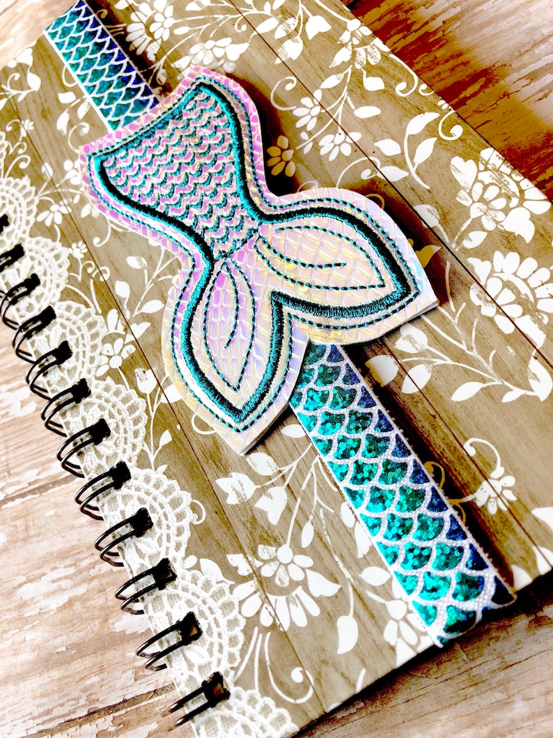 Puede incluir: Un cuaderno con un marcap&aacute;ginas en forma de cola de sirena. El marcap&aacute;ginas est&aacute; hecho de tela iridiscente con un esquema de color azul y verde. El cuaderno tiene un dise&ntilde;o floral marr&oacute;n y blanco.