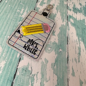 May include: A silver glitter keychain with a pencil design and the text "Mrs. White" on a white background with blue lines.