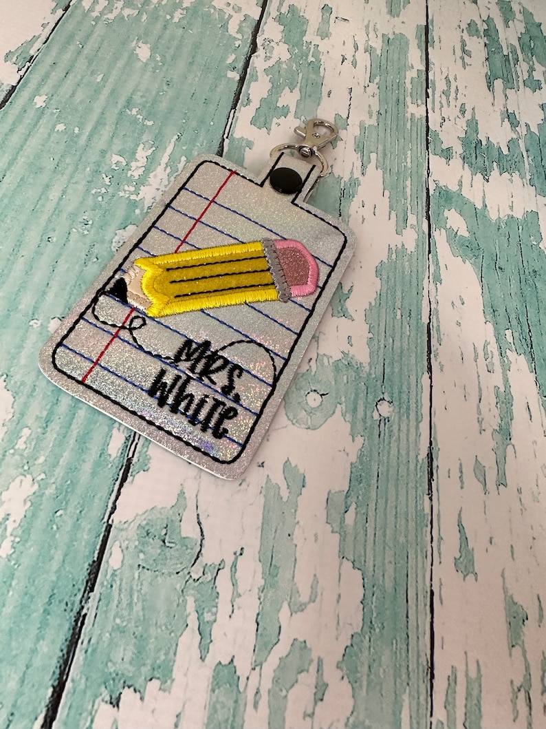 May include: A silver glitter keychain with a pencil design and the text "Mrs. White" on a white background with blue lines.