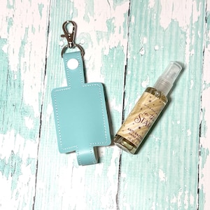 May include: A light blue leather hand sanitizer holder with a silver keychain and a white button. The holder is next to a small clear bottle of hand sanitizer with a gold label that says "Bath & Body Works In the Stars Anti-Bacterial Hand Spray 1 fl oz".