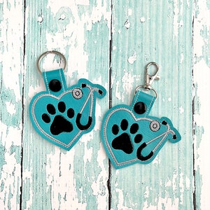 May include: Two teal blue heart-shaped keychains with a black paw print and a silver stethoscope. The keychains are on a white wooden background.