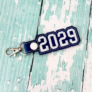 May include: A navy blue and white keychain with the year "2029" embroidered on it. The keychain has a silver clasp and a navy blue strap. The design is in a collegiate style.