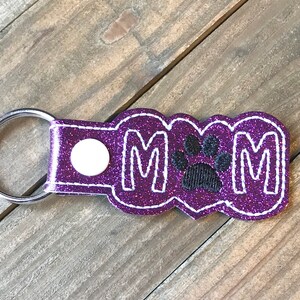 May include: Purple glitter keychain with the letters "MOM" and a black paw print in the center.