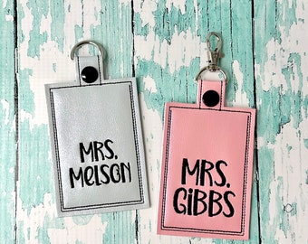May include: Two keychains with silver and pink fabric, each with a black button and a silver ring. The silver keychain has the text "MRS. MELSON" and the pink keychain has the text "MRS. GIBBS".