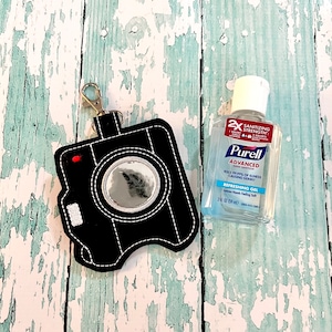 May include: Black camera-shaped keychain with a small mirror inside and a silver clip. Next to it is a clear plastic bottle of Purell Advanced Hand Sanitizer with the text "2x Sanitizing Strength" and "Refreshing Gel" on the label.