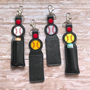 May include: Four black leather keychains with a softball design. Each keychain has a different colored softball with white stitching. The keychains have a silver clip and a red loop.