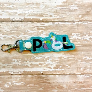 May include: A blue felt keychain with the word "POOL" spelled out in black felt letters. The letters are decorated with embroidered images of a beach ball, a unicorn float, and a pool noodle.
