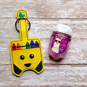 May include: A yellow crayon box-shaped hand sanitiser holder with a key ring. The holder has a smiling face and a row of colourful crayons. A pink hand sanitiser bottle with a llama wearing sunglasses and the text "No Llama Drama" is next to the holder.