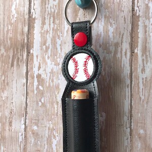 May include: Black leather keychain with a red button and a white baseball with red stitching. The keychain is designed to hold a small tube of essential oil or lip balm.