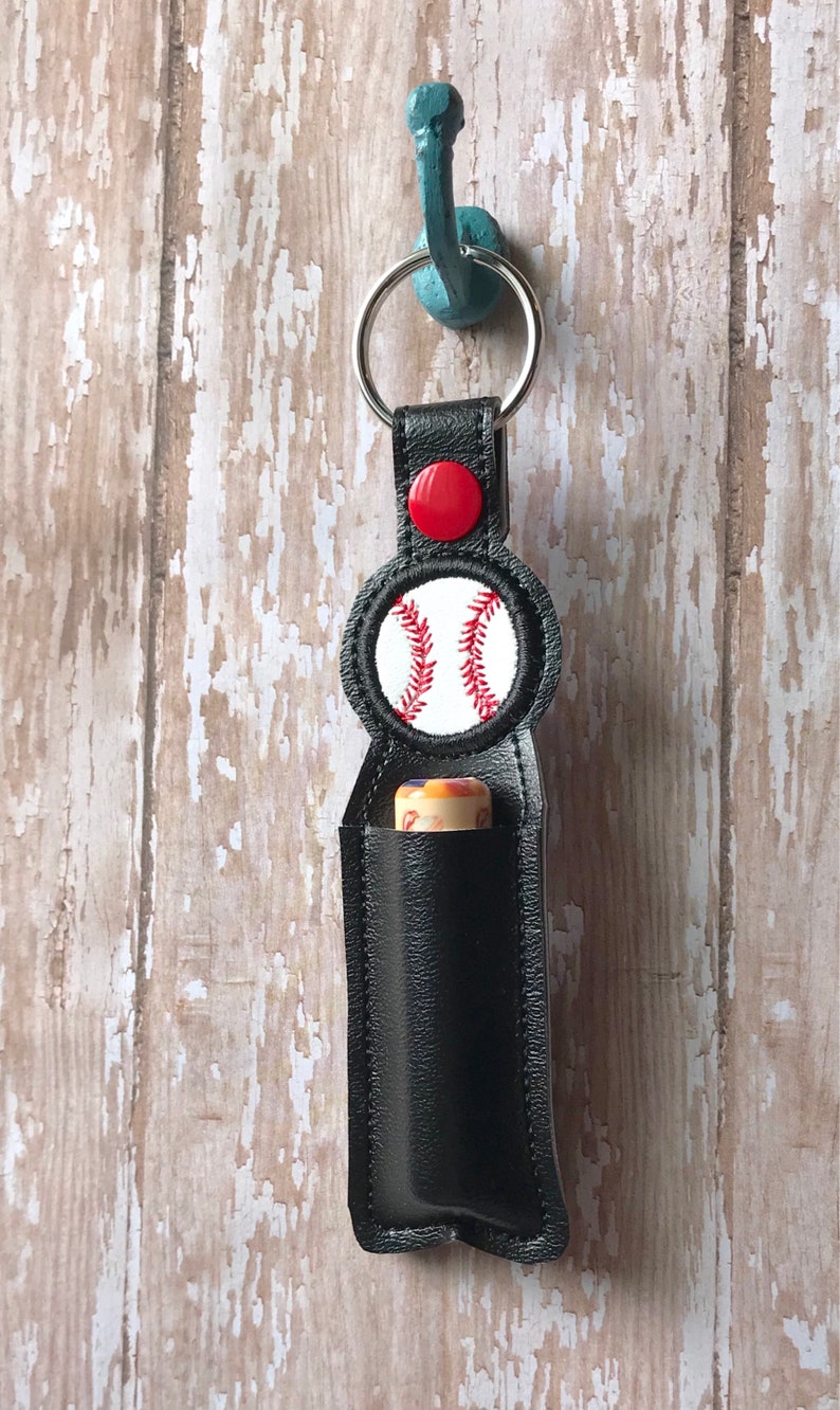 May include: Black leather keychain with a red button and a white baseball with red stitching. The keychain is designed to hold a small tube of essential oil or lip balm.