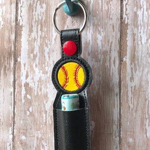 May include: Black leather keychain with a softball design and a red button. The keychain has a pocket for a lip balm or other small item.