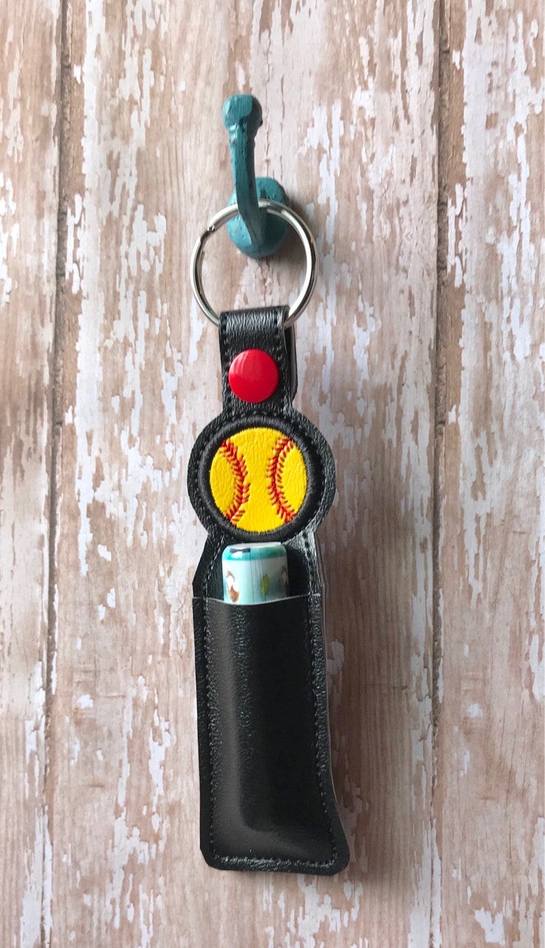 May include: Black leather keychain with a softball design and a red button. The keychain has a pocket for a lip balm or other small item.