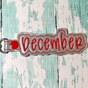 May include: Silver glitter keychain with a red button and the word "December" embroidered in red thread.