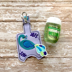 May include: Purple fabric hand sanitizer holder with a peacock design and a silver keychain. The holder is attached to a clear plastic bottle of hand sanitizer labeled "PocketBac Bath & Body Works Cucumber Melon Anti-Bacterial Hand Gel 1 fl oz / 29 ml."