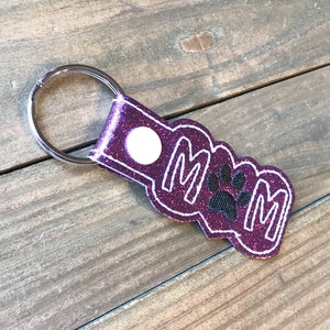 May include: Purple glitter keychain with the word "MOM" and a black paw print in the center. The keychain has a silver metal ring.