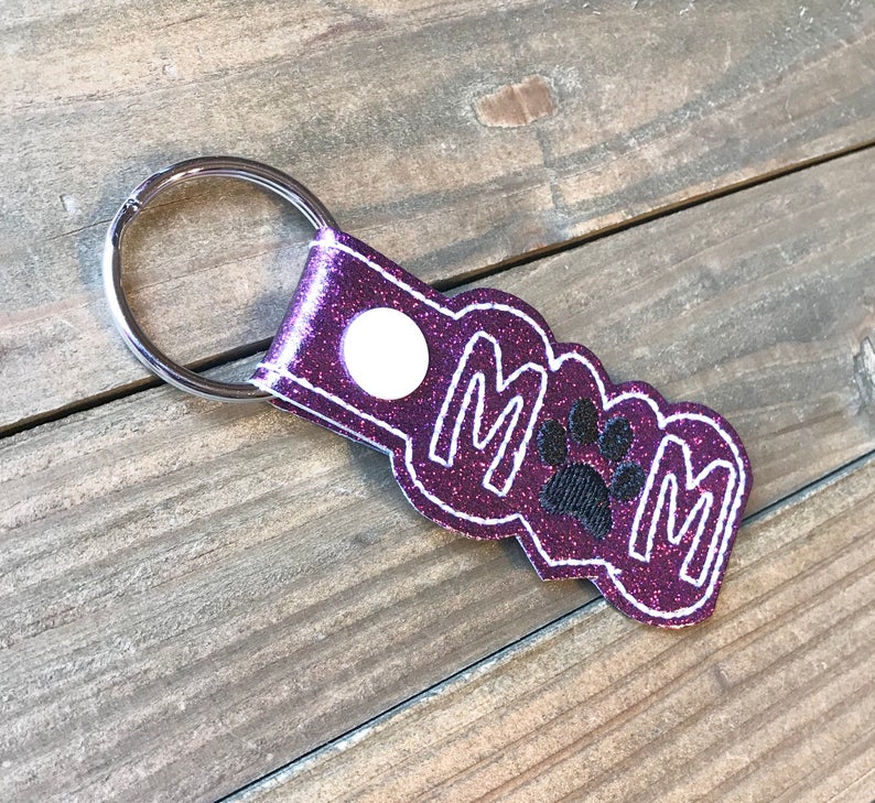 May include: Purple glitter keychain with the word "MOM" and a black paw print in the center. The keychain has a silver metal ring.