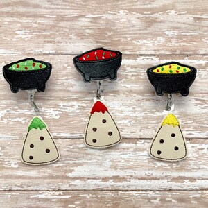 May include: Four embroidered badge reels featuring a variety of Mexican food designs. The designs include a bowl of guacamole, a bowl of salsa, a nacho with red salsa, and a nacho with cheese.