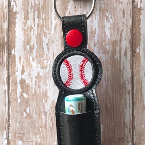 May include: Black leather lip balm holder with a red button and a white baseball with red stitching. The holder is attached to a silver key ring.