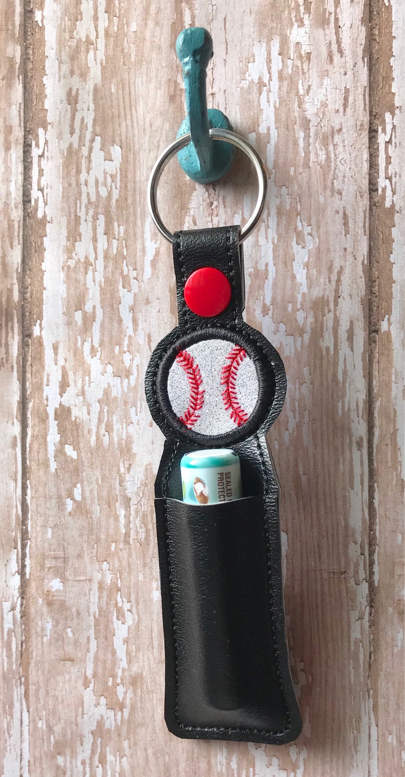 May include: Black leather lip balm holder with a red button and a white baseball with red stitching. The holder is attached to a silver key ring.