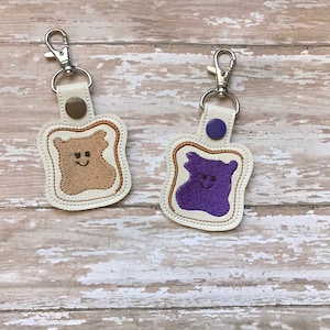 May include: Two keychains shaped like slices of bread with embroidered faces. One slice has brown peanut butter filling and the other has purple jelly filling.