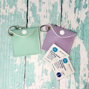 May include: Two small, square pouches in mint green and lavender, each with a silver keyring and a white snap closure. Two sterile alcohol prep pads are next to the pouches. The pouches are on a distressed, light blue and white wooden surface.