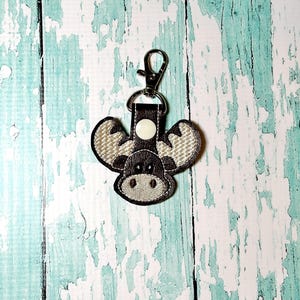 May include: A brown moose keychain with a white button and a silver clip. The moose has a white face and antlers.
