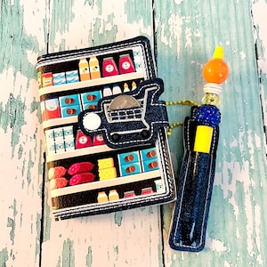 May include: A blue denim wallet with a grocery store aisle print and a shopping cart charm. A blue denim pen holder with a yellow top and a beaded charm is attached to the wallet.