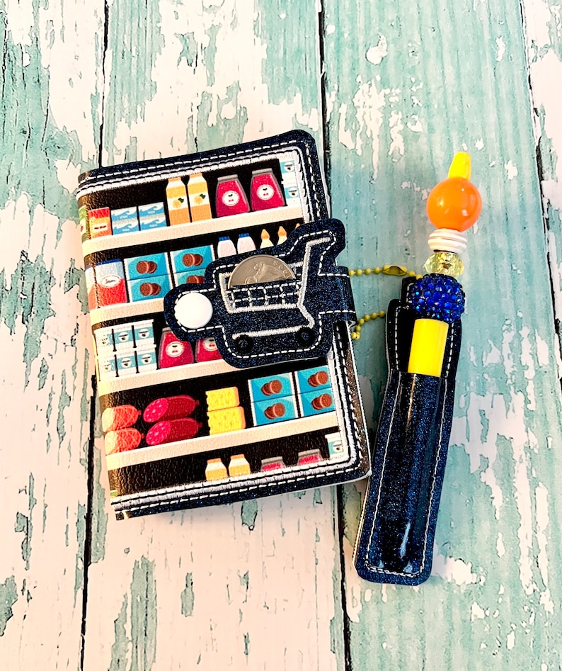 May include: A blue denim wallet with a grocery store aisle print and a shopping cart charm. A blue denim pen holder with a yellow top and a beaded charm is attached to the wallet.