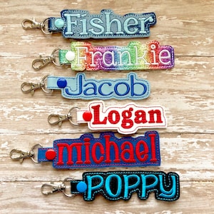 Pode incluir: Seis chaveiros personalizados com diferentes nomes bordados. Os nomes s&atilde;o Fisher, Frankie, Jacob, Logan, Michael e Poppy. Cada chaveiro tem uma cor e um design diferente.